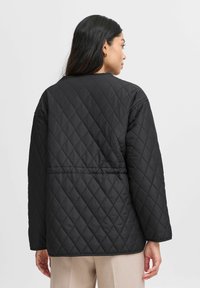 Schwarze gesteppte Jacke mit lockerer Passform, rundem Kragen und langen Ärmeln. Das strukturierte Muster besteht aus Rautenformen.