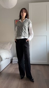 Szürke kötött mellény kábelmintával, fehér galléros ing fölött, fekete bő szárú nadrággal és fekete hegyes orrú magassarkúval párosítva.