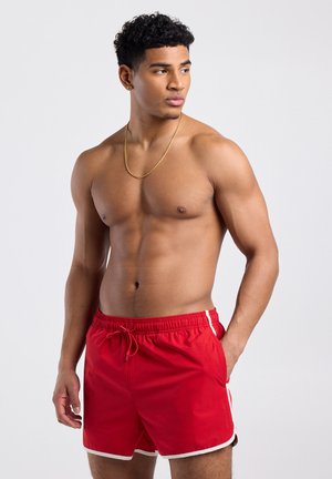 Gespierde man met kort krullend haar, gekleed in rode zwemshorts met witte rand, een gouden ketting en oorbellen, kijkt naar links tegen een effen achtergrond.