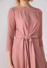 Rosa 3/4 ermers kjole i et teksturert stoff. Har et vridd knutedetalj i midjen og en rund hals. Glatt finish, avslappet passform.