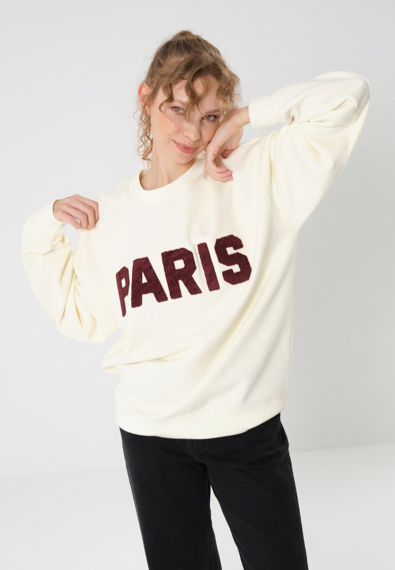 Felpa crema con la scritta "PARIS" in lettering borgogna testurizzato, colletto rotondo e spalle scese, abbinata a pantaloni neri.