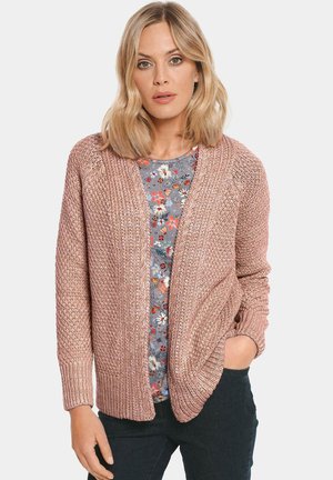 Strickjacke in sanftem Rosa mit offener Vorderseite, gerippten Bündchen und strukturierten Mustern. Getragen über einem blumendrucktop und dunklen Hosen.