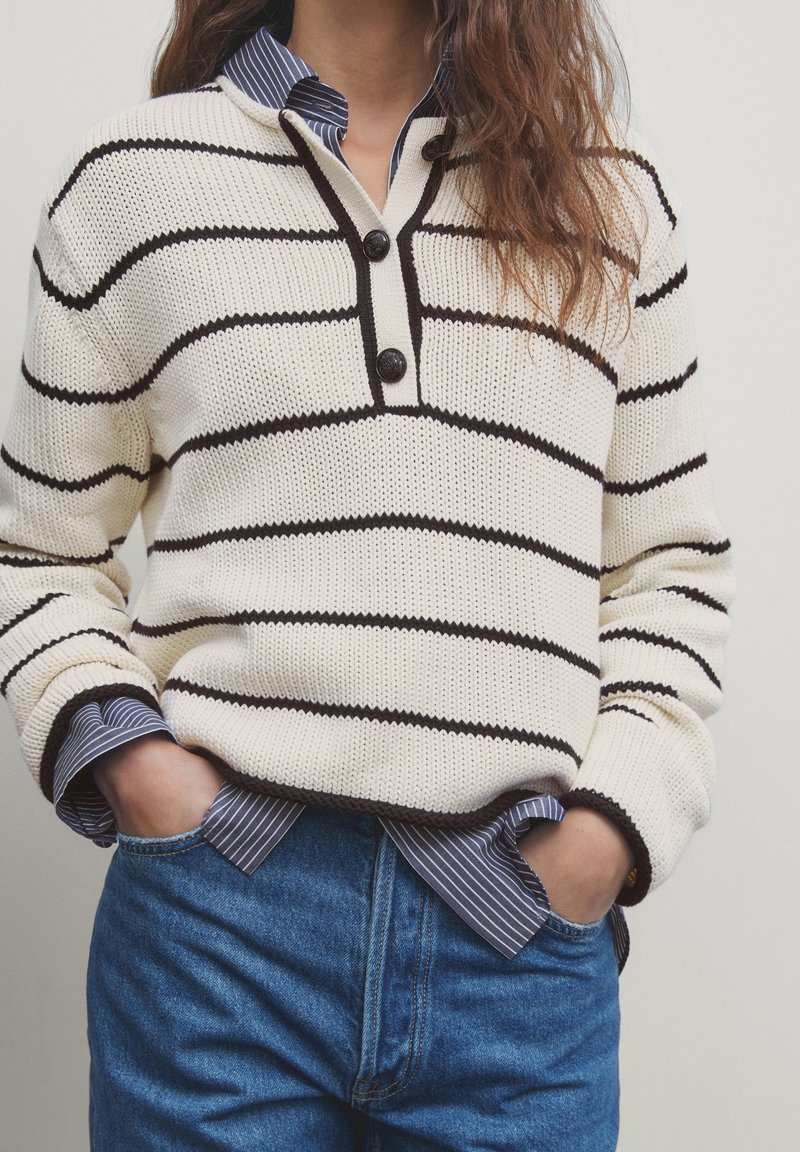 Personne portant un pull en tricot crème avec des rayures horizontales noires, deux boutons, superposé sur une chemise rayée bleue et blanche, mains dans les poches d’un jean bleu.