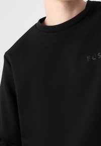 Zbliżenie na osobę w czarnej bluzie z okrągłym dekoltem z subtelnym logo "BOSS" na piersi, na jednolitym tle.