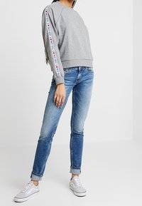 Sudadera gris con dobladillos y puños acanalados, que presenta una franja multicolor con "I ❤️ NY" en la manga izquierda. Combinada con jeans ajustados azules y zapatillas grises.
