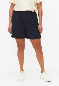 Pantaloni corti blu navy con un motivo testurizzato, vita elastica e bordo in pizzo, abbinati a sneakers bianche.