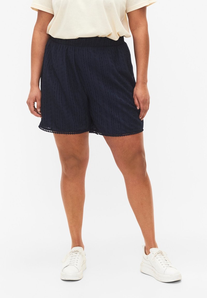 Pantaloni corti blu navy con un motivo testurizzato, vita elastica e bordo in pizzo, abbinati a sneakers bianche.