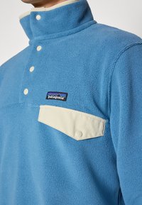 Fleece pullover i lys blå med høj krave, med en cremefarvet lommeklap og Patagonia-logo broderet på brystet.