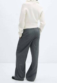 Femme portant un pull col montant crème et un pantalon gris à jambe large, debout avec les mains dans les poches, de dos.