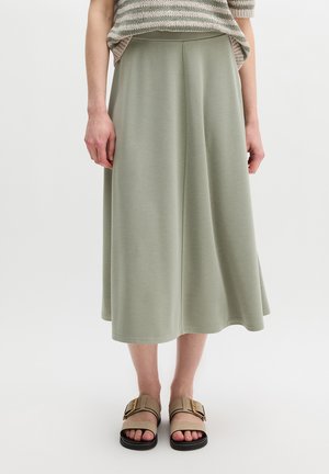 Vrouw draagt een midirok in saliegroen, een beige gestreept shirt met korte mouwen en beige sandalen met gesp, staand op een witte vloer.