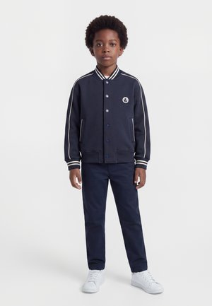 Garçon debout, portant une veste varsity bleu marine avec des bandes blanches, un pantalon assorti et des baskets blanches sur un fond clair uni.