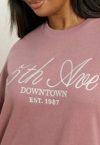 Vaaleanpunainen huppari, jossa on valkoisella brodeerattu teksti "5th Ave DOWNTOWN EST. 1987." Yksinkertainen pyöreä kaula-aukko ja pehmeä rakenne.