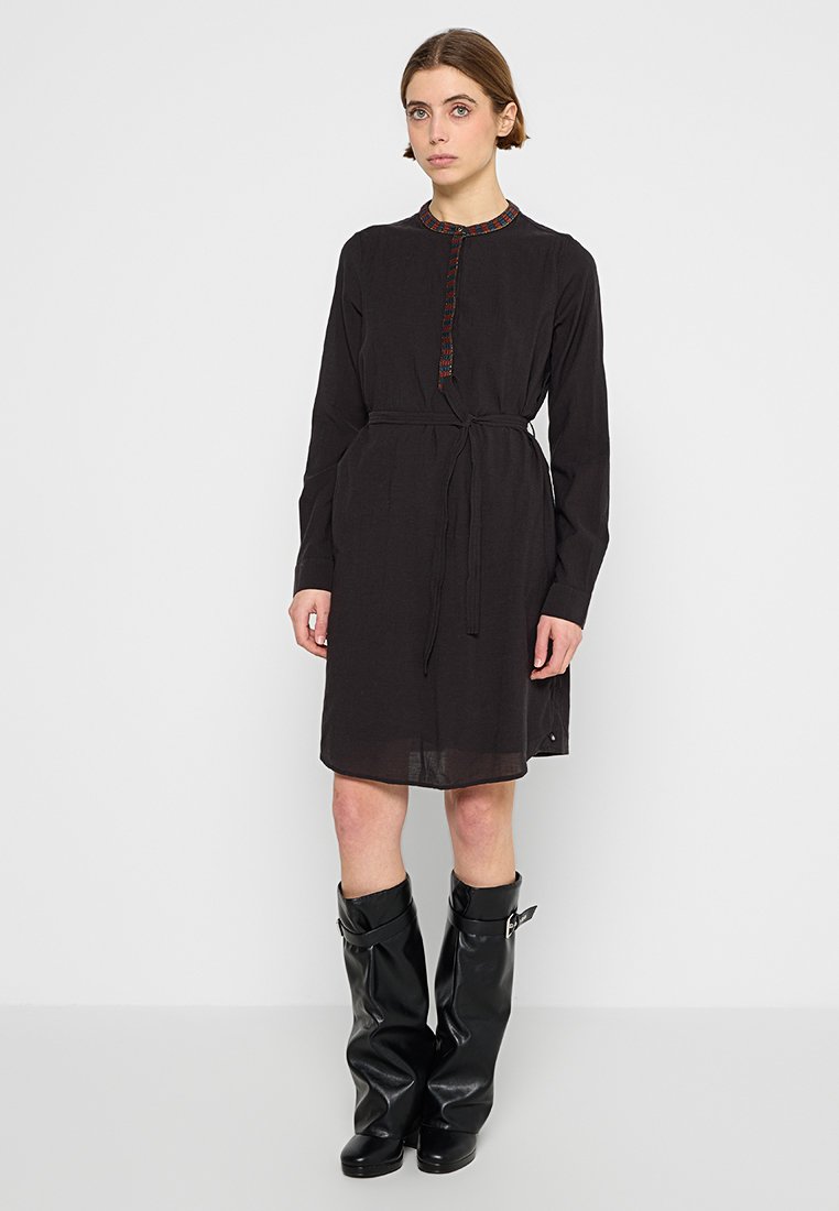 Schwarzes langärmliges Kleid mit Bindegurt, strukturiertem Stoff und dekorativen Knöpfen entlang des Ausschnitts. Knielang mit geradem Schnitt.