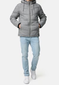 Mies, który ma na sobie szary ocieplany płaszcz z kapturem, jasnoniebieskie dżinsy i białe trampki, trzyma ręce w kieszeniach płaszcza, stojąc na białym tle.