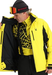 Gelbe und schwarze isolierte Skijacke mit einem Frontreißverschluss, internem Mesh-Design und Reißverschlusstaschen. Verfügt über verstellbare Bündchen und eine warme Fütterung.