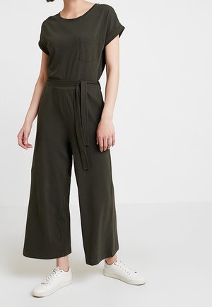 Femme portant une combinaison courte à manches courtes vert olive foncé avec ceinture à la taille et des baskets blanches, se tenant devant un fond clair uni.