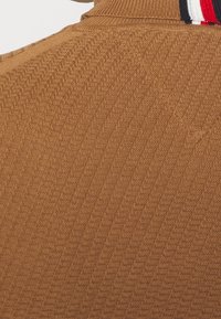 Pull en tricot marron texturé avec un col roulé côtelé et un petit détail vertical à rayures rouges, blanches et noires près du col.