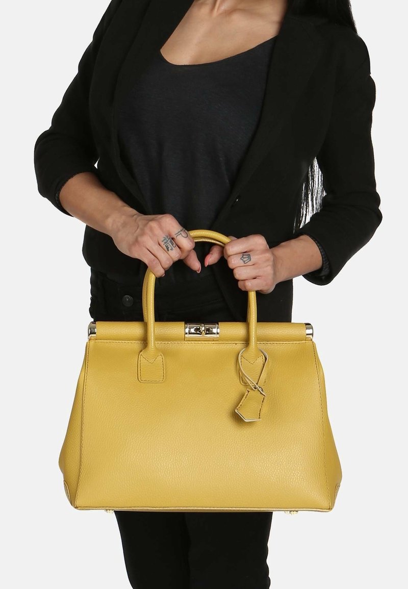 Femme en noir tenant un grand sac à main en cuir jaune avec des poignées supérieures et une petite étiquette en cuir attachée.