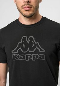 Sort bomulds t-shirt med et stort, mørkegrå Kappa-logo, hvor to silhuetfigurer sidder ryg-til-ryg. Rund halsudskæring.