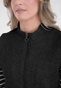 Zwarte geweven jas met een hoge kraag en rits, met gestreepte stof op de binnenmouwen. Het gestructureerde materiaal contrasteert met gladde accenten.