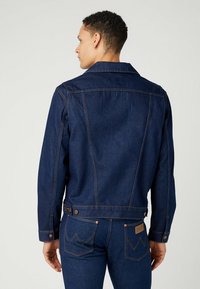 Chaqueta de mezclilla en azul oscuro con un cuello clásico, cierre de botones y costuras visibles. Cuenta con bolsillos en la parte trasera y los lados.
