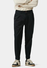 Zwarte geplooide broek met een relaxte fit, knoopsluiting en opgerolde mouwen, gecombineerd met casual zwart-witte sneakers.