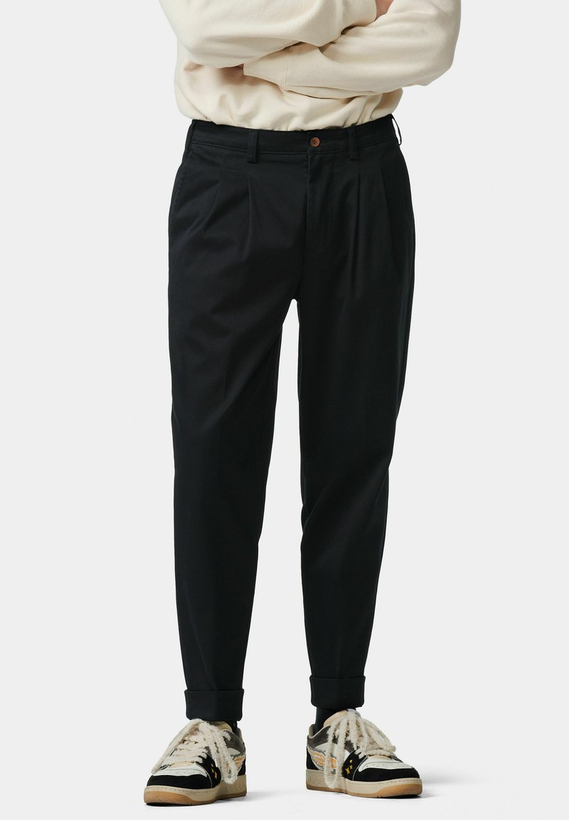 Zwarte geplooide broek met een relaxte fit, knoopsluiting en opgerolde mouwen, gecombineerd met casual zwart-witte sneakers.