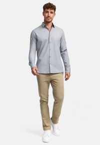 Camisa de botones gris claro con mangas largas, combinada con pantalones caqui y zapatillas blancas, de pie sobre un fondo neutro.