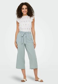 Blouse blanche avec bordure en dentelle et détails ajourés, associée à un pantalon large vert clair et des sandales blanches.