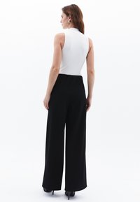 Pantaloni neri a gamba larga abbinati a un top bianco aderente senza maniche, caratterizzato da un colletto alto e una texture liscia. Tacchi neri completano il look.