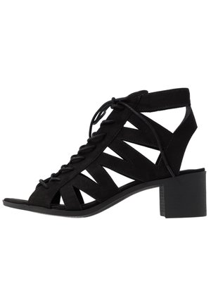 Czarna zamszowa sandał na obcasie z odkrytym noskiem, wiązaniem na przodzie i wycięciami po bokach. Średni obcas typu klocek z teksturowanym wykończeniem.