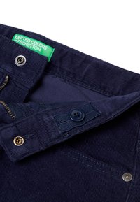 Granatowe spodnie z korduroju z zamkiem, zapięciem na guzik i złotymi dodatkami. Zielona metka z napisem "United Colors of Benetton." Gładka tekstura.