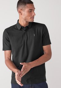 Camisa polo negra de algodón con cuello, mangas cortas y un pequeño logo bordado en el pecho. Textura suave, corte clásico.