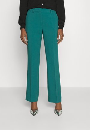 Pantalon large couleur sarcelle avec une texture lisse, présentant une coupe ajustée et un pli avant. Associé à des talons métalliques à bout pointu.