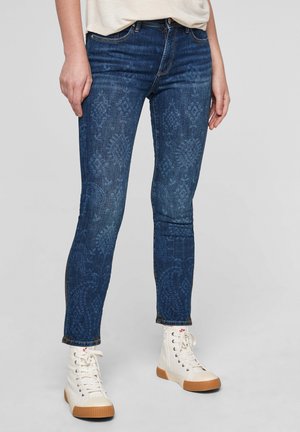 Jean skinny en denim bleu foncé avec une texture à motif subtil. Comprend un design classique à cinq poches et un ourlet au niveau de la cheville, associé à des baskets blanches montantes.