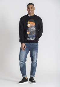 Sudadera negra con un oso de dibujos animados que lleva una camiseta gris y jeans azules, combinado con denim desgastado y zapatillas negras.