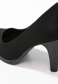 Chaussure à talon haut en suède noir avec un talon carré texturé et pailleté sur fond blanc, montrant les détails latéraux et inférieurs.