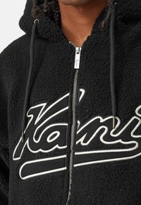 Felpa nera con zip e cappuccio foderato, zip argentata e grande scritta bianca 'Kani' sul davanti. Materiale morbido e texture piacevole.