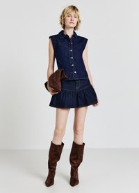 Abito camicia senza maniche in denim scuro, con bottoni sul davanti e gonna a campana, abbinato a stivali al ginocchio in suede marrone, mentre tiene un tessuto in una mano.