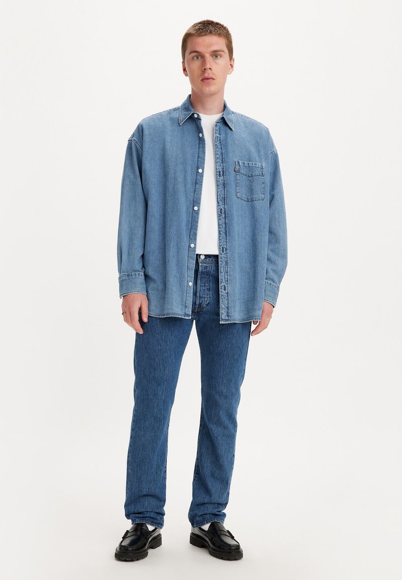 Chemise en denim bleu clair, design à boutons avec une poche poitrine. Associée à un jean bleu et des chaussures noires. Le modèle se tient devant un fond blanc.