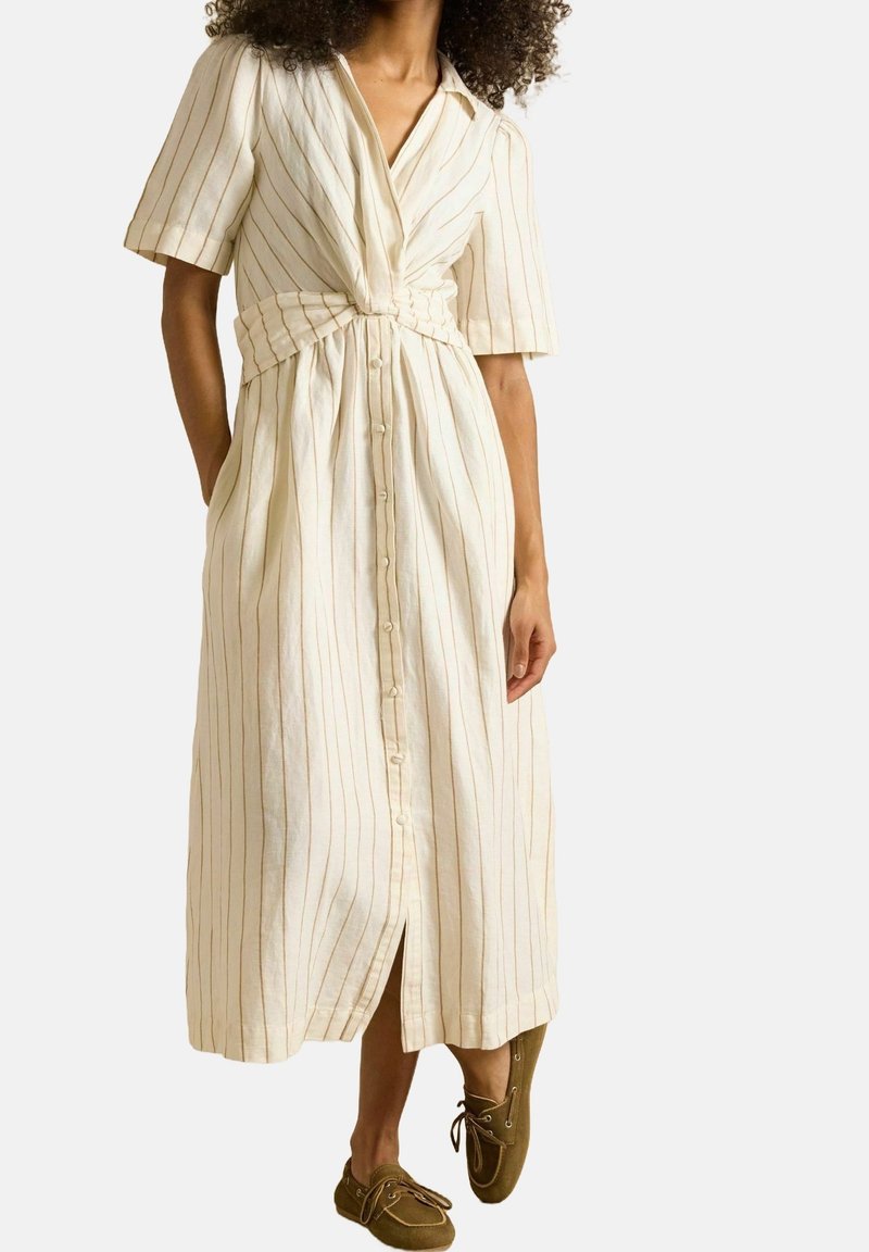 Femme portant une robe midi crème à rayures verticales, avec des boutons sur le devant et des manches courtes, associée à des chaussures à lacets vert olive.