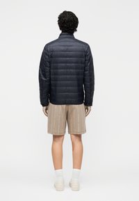 Geacă matlasată de culoare bleumarin, cu model matlasat, guler înalt și mâneci lungi, asortată cu pantaloni scurți bej cu un model repetat de logo.