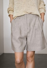 Maglione lavorato a maglia beige e pantaloni shorts plissettati grigio chiaro. I pantaloni shorts presentano tasche laterali e un sottile motivo testurizzato.