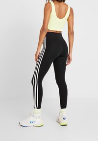 Kvinna som bär svarta leggings med vita sidostråkar, en gul croppad linne, randiga strumpor och vita sneakers stående på ett helt vitt golv.