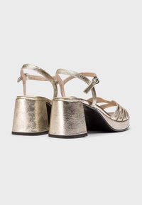 Sandales à talons bloc métalliques dorées avec des brides entrecroisées, bout ouvert et dessus texturé. Comprend une bride ajustable à la cheville pour un meilleur maintien.