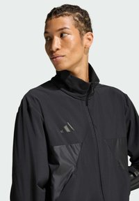 Crna atletska jakna s visokim ovratnikom, zatvaranjem na dupli zip i kontrastnim sjajnim panelima. Ima logo na prsima i koristi laganu tkaninu.