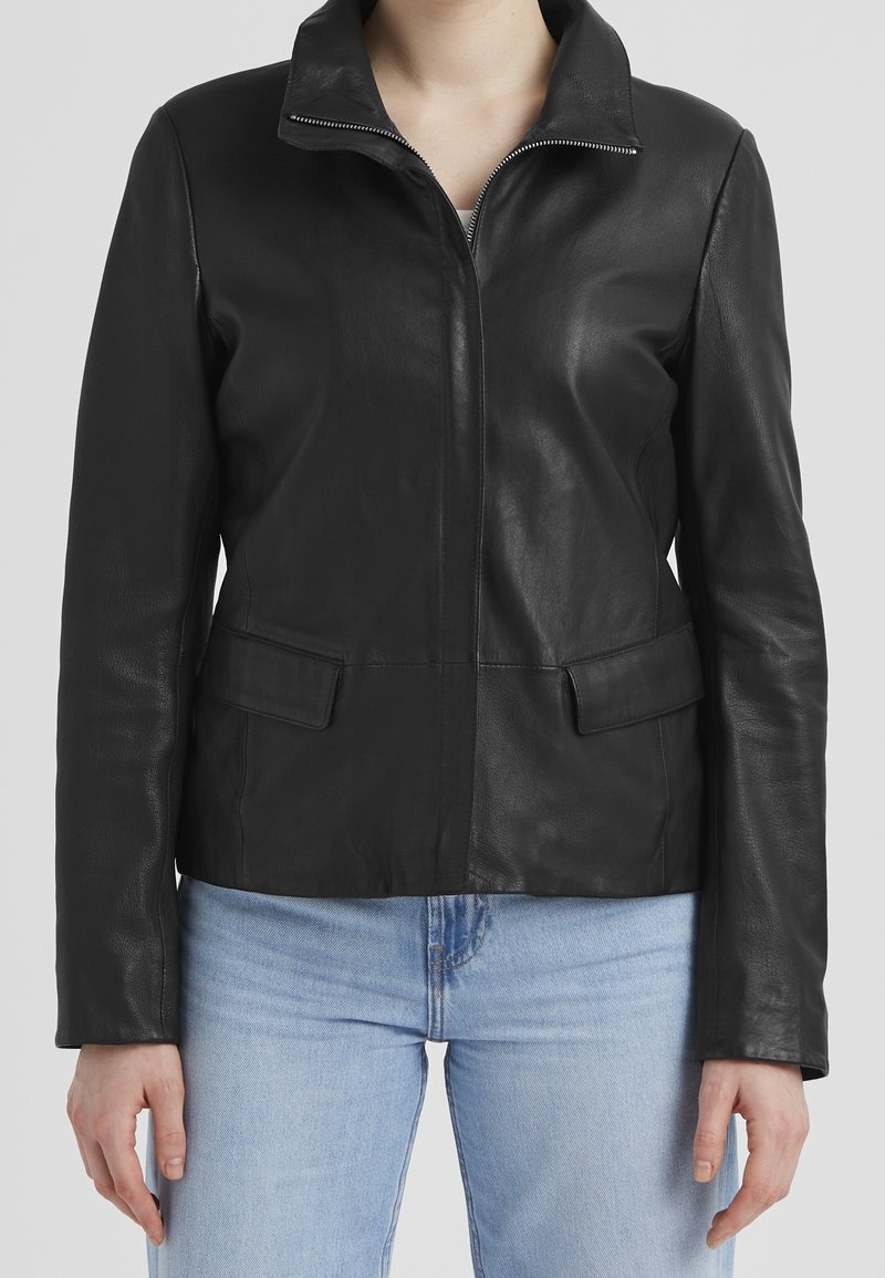 Žena v dobře padnoucí černé kožené bundě na zip s klopovými kapsami a světle modrých denimech, stojící na jednobarevném pozadí.