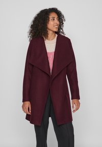 Burgundijas vilnas mētelis ar plandošu apkakli, garām piedurknēm un taisnu piegriezumu; virsū uzvilkts gaišs svītrains adījums.