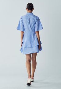 Blau gestreiftes Hemdkleid mit kurzen Ärmeln, tailliertem Schnitt und geschichtetem Saum. Worn mit weißen, hohen Sandalen. Rückansicht wird gezeigt.