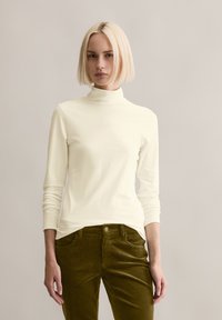 Cremfarbener Rollkragenpullover mit langen Ärmeln aus weichem Stoff, kombiniert mit olivgrünen Cordhosen. Verfügt über ein tailliertes Design und gerippte Textur.